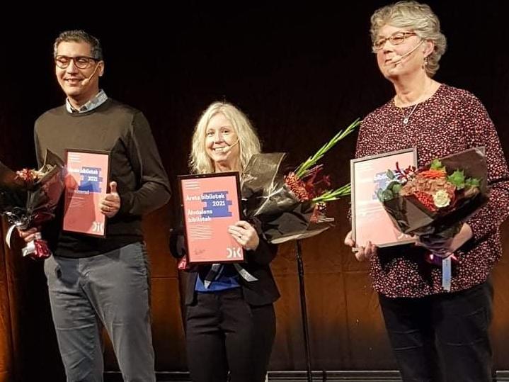 Tre glada personer med diplom och blommor i händerna. Utdelning av priset Årets bibliotek på Bokmässan i Göteborg.