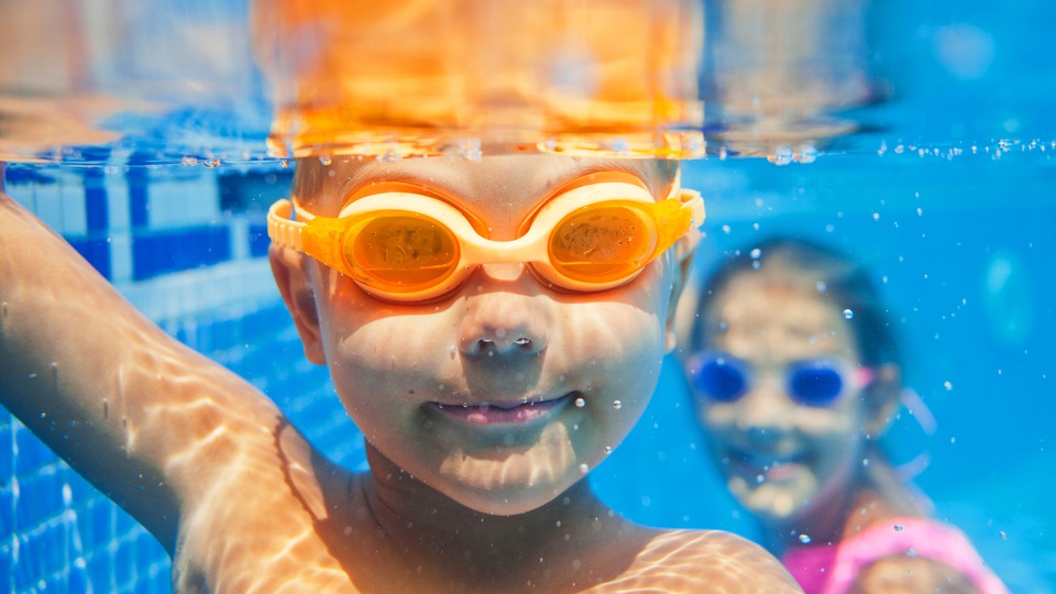 Två barn med simglasögon under vatten i bassäng. 