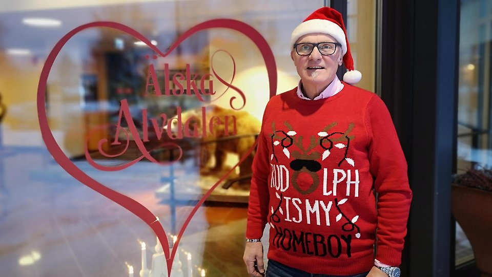 En man iklädd röd jultröja och tomteluva står framför ett stort fönster där man i ett hjärta kan läsa Älska Älvdalen.