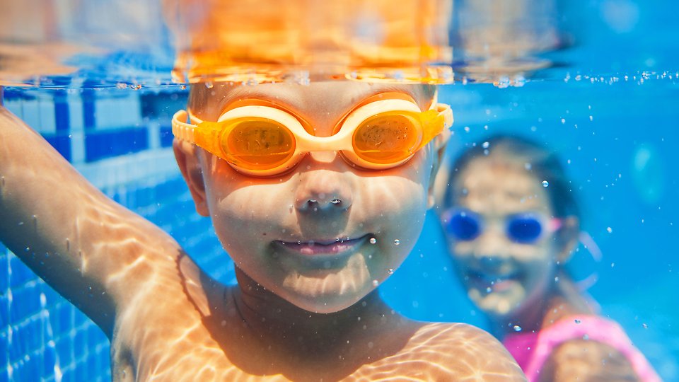 Två barn med simglasögon under vattnet i simbassäng.