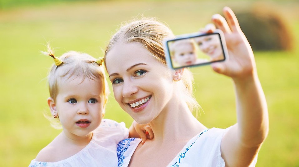 En mamma med sin dotter på armen tar en selfie i ett sommarlandskap