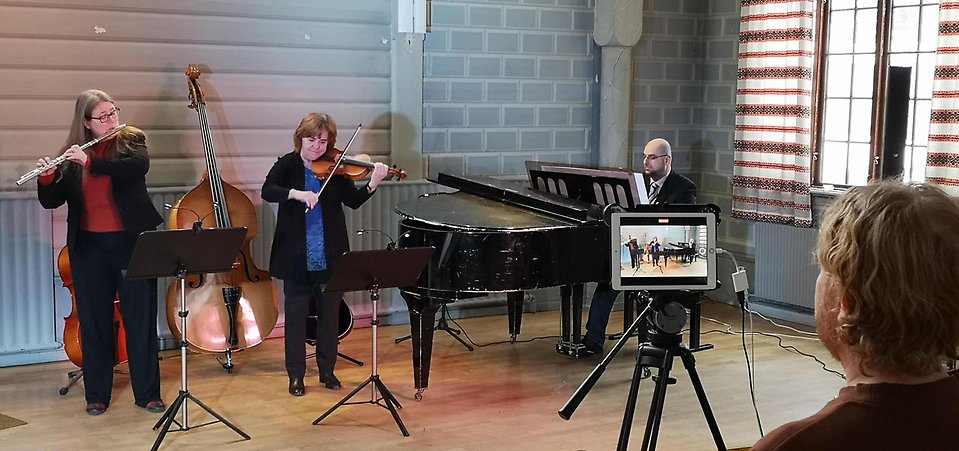 En flöjtist, en violinist och en pianist spelar ett stycke musik på en scen. De spelas in på en iPad av en man.