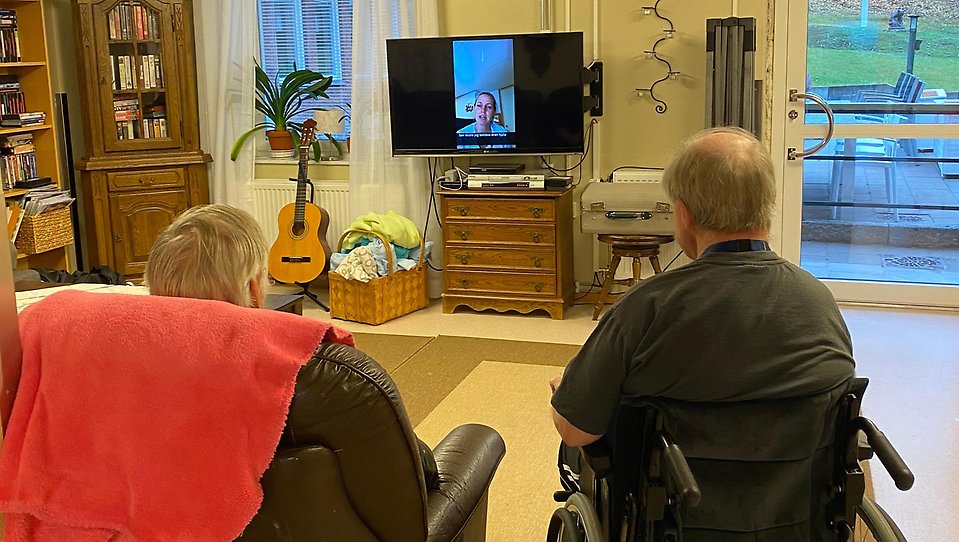 Två personer på ett äldreboende sitter framför en TV och tittar på en inspelad videohälsning